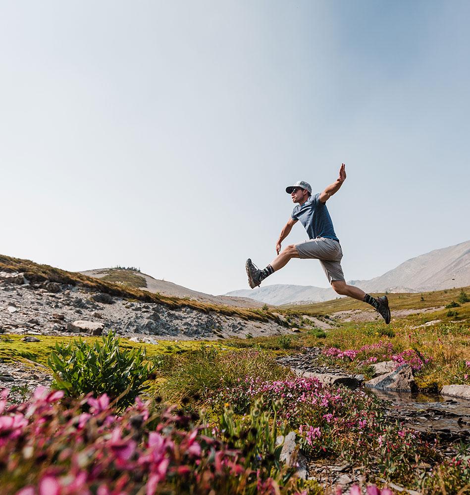 Man wearing a cap and hiking gear, leaping over a small stream surrounded by wildflowers in a mountainous landscape on a clear, sunny day.
