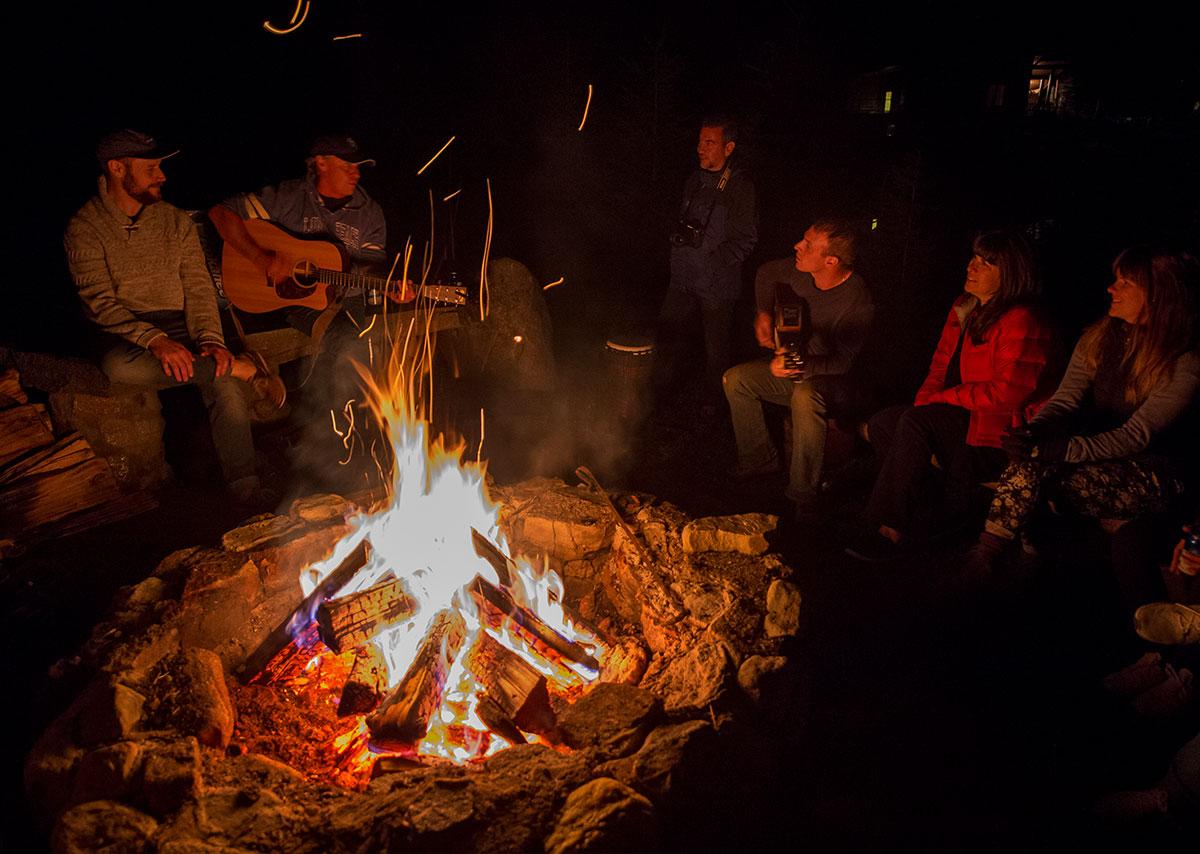 Group of people sitting around a campfire, with two individuals playing guitars.