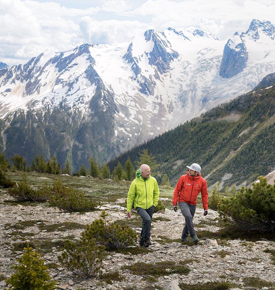 Two male hikers walking and talking in a mountainous landscape with snow-covered peaks in the background.