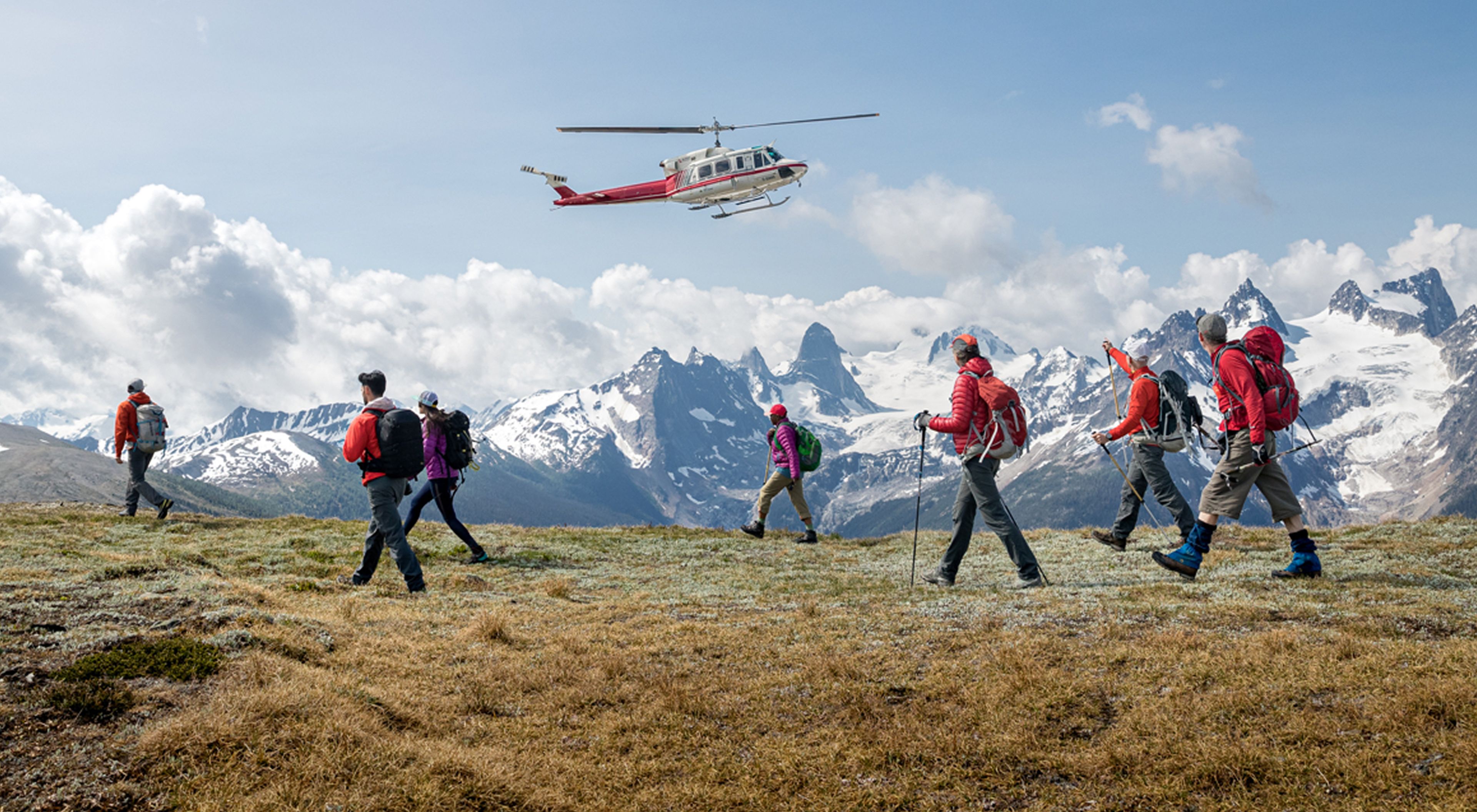 A group of hikers with backpacks walks across a grassy alpine ridge while a red-and-white helicopter flies low overhead. In the foreground, several people use trekking poles and wear outdoor gear in bright colors. 
