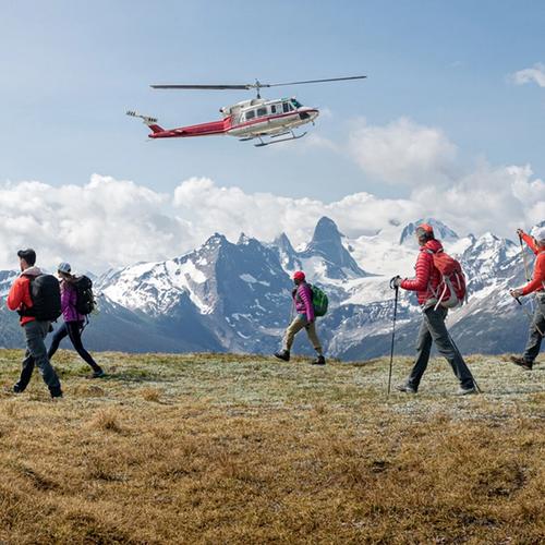 A group of hikers with backpacks walks across a grassy alpine ridge while a red-and-white helicopter flies low overhead. In the foreground, several people use trekking poles and wear outdoor gear in bright colors.