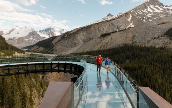 two people walking on a glass bridge high over trees with mountains in the background