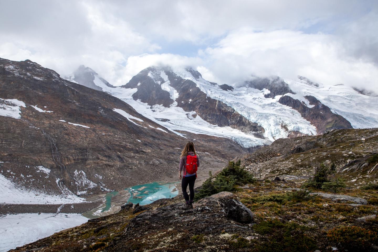A woman with a red backpack on a ridge looking at a glacier