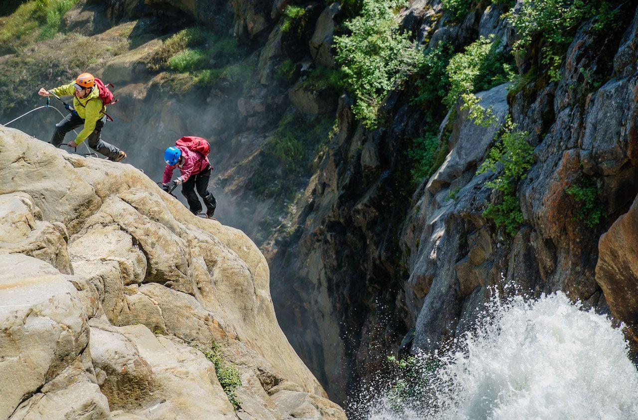 Two individuals wearing a helmet and harness climb up the side of a rock face on a CMH via ferrata. 