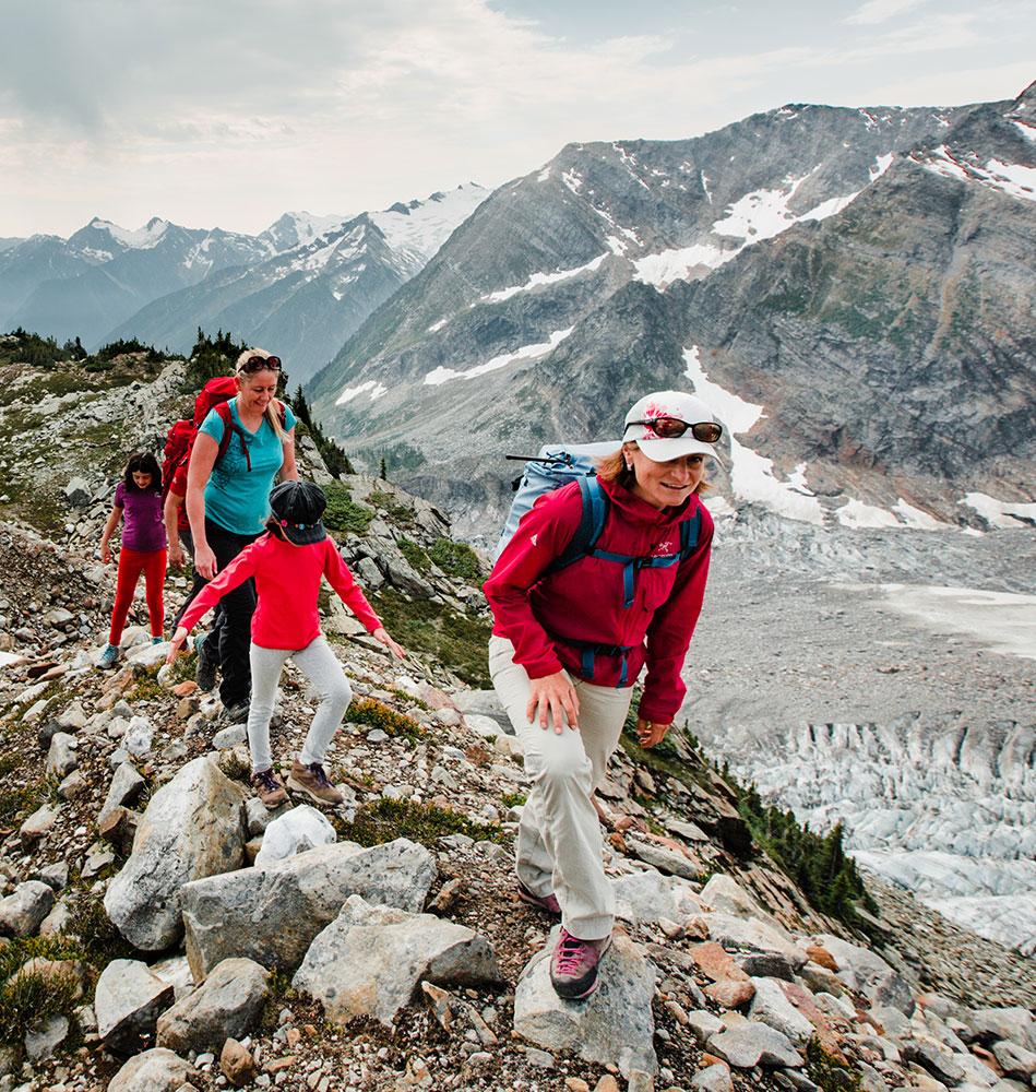 Group of hikers, including children and adults, climbing a rocky mountain trail with glacier views and snow-capped peaks in the background