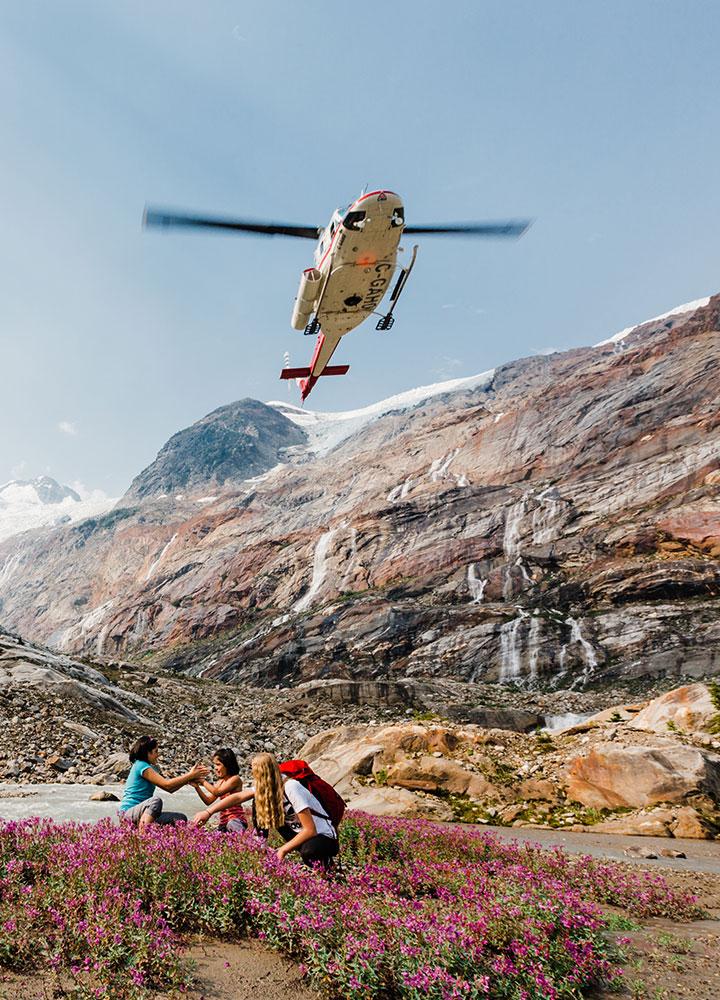 Helicopter flying overhead as three hikers sit among wildflowers in a mountain valley with rocky cliffs and waterfalls in the background