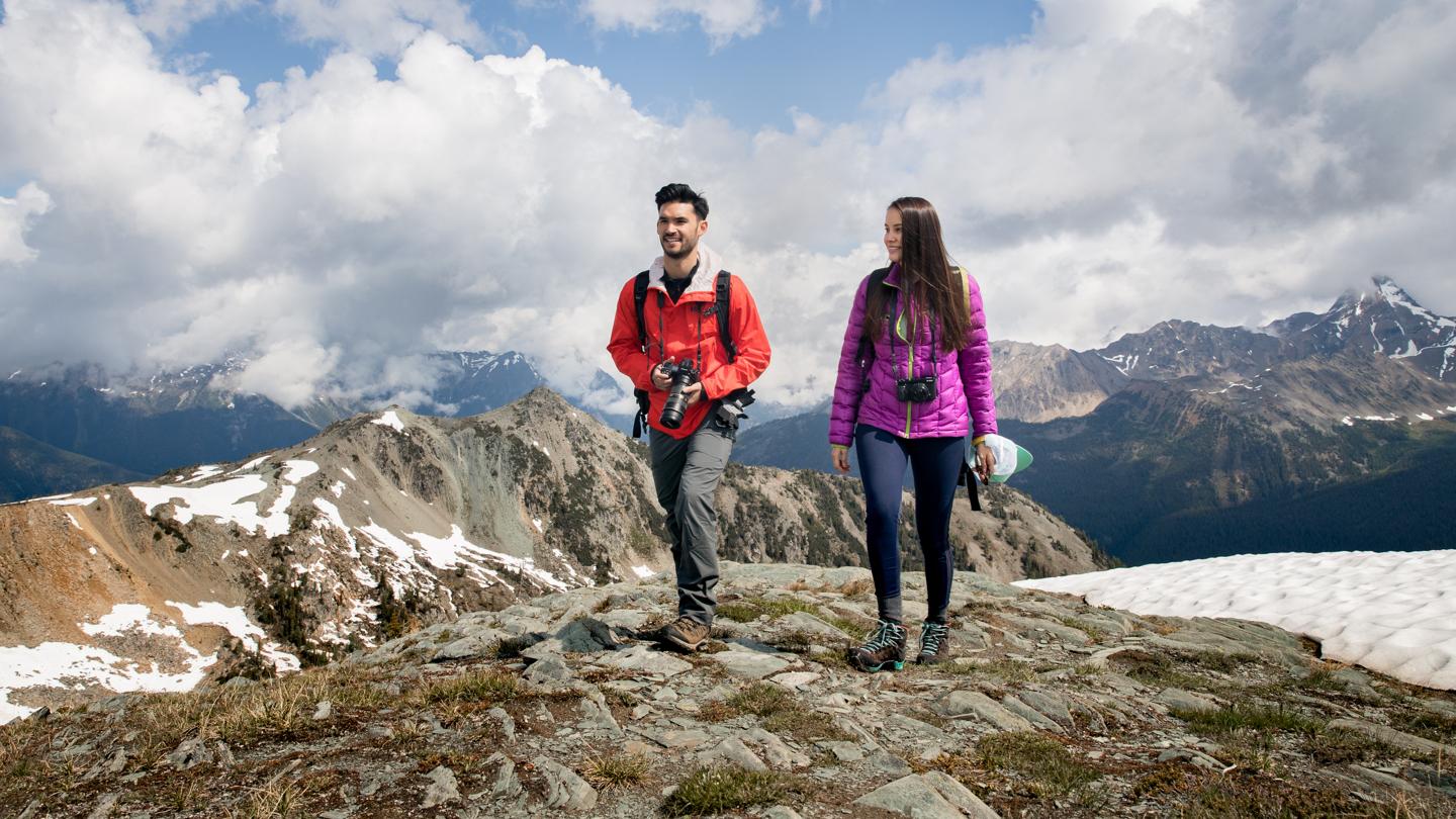 two people walking on a ridge with cameras and mountain in the background