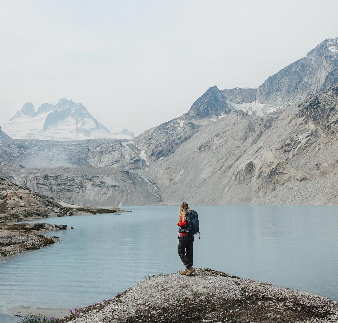 A hiker wearing a backpack stops at a view point overlooking a clear blue lake with jagged peaks rising in the background.