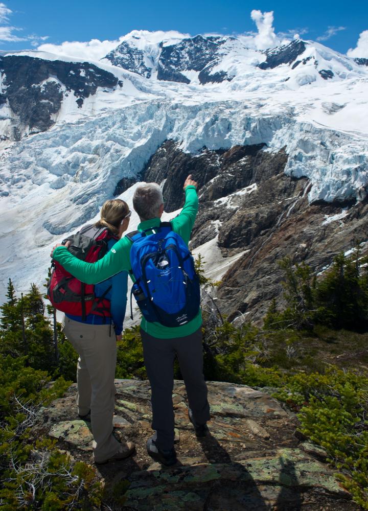 two people looking out from a ridge at a glacier, one of the men has his arm around the other and is pointing to the glacier