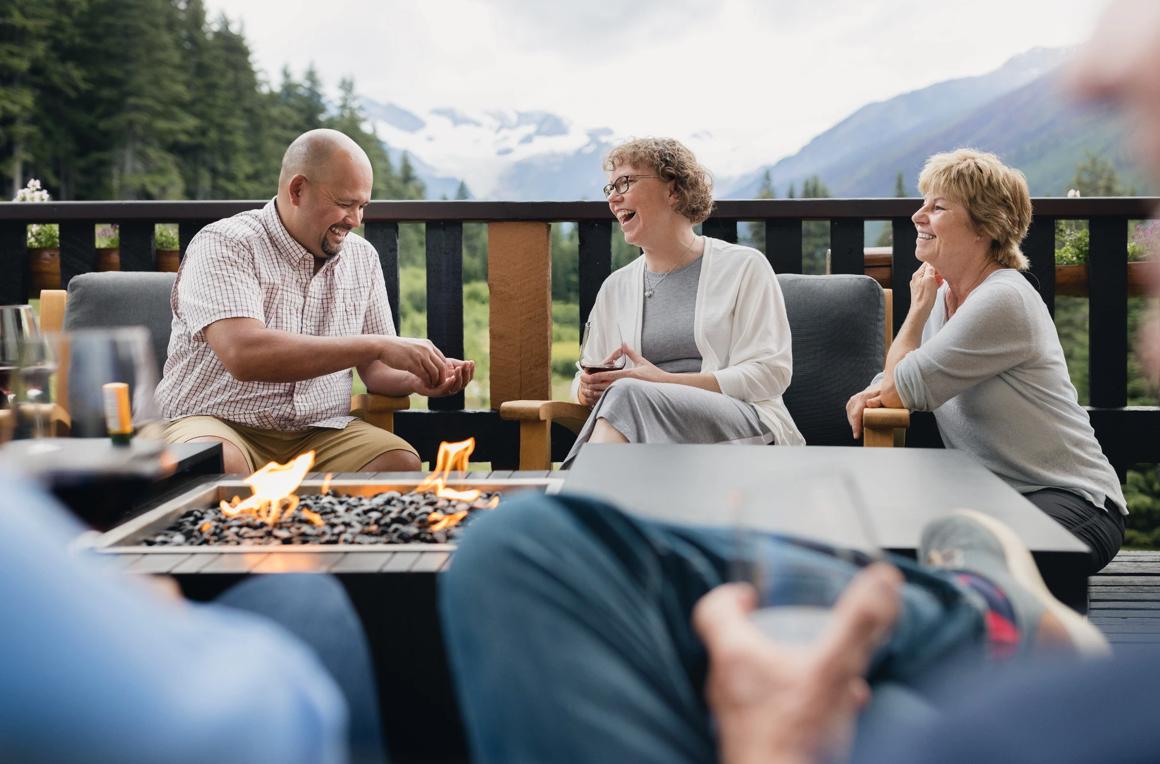 People sitting around a fire table at a lodge laughing with mountains in the background