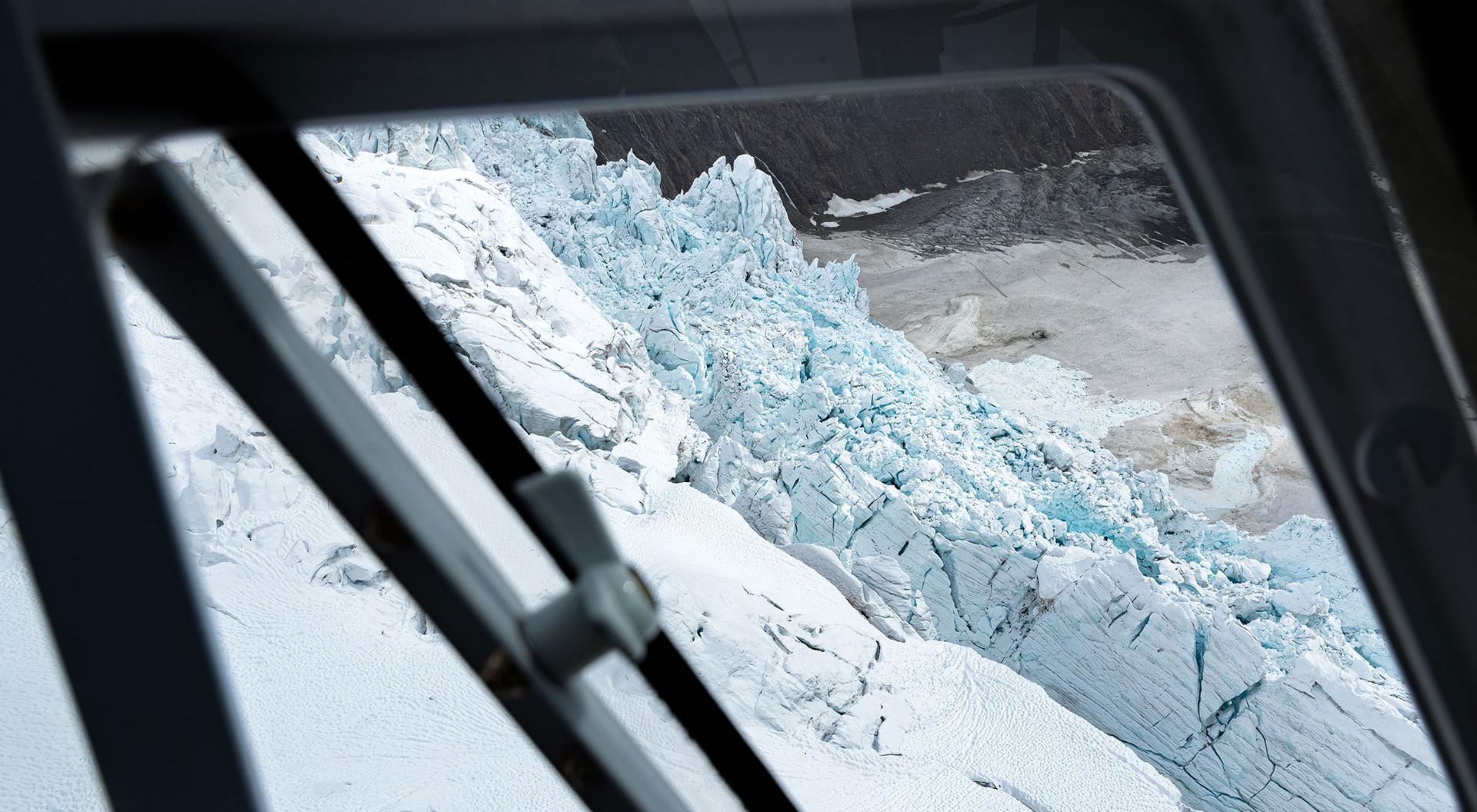 An aerial top-down view looking through a helicopter window at a massive, crevassed glacier and snow field during a CMH Summer Adventures heli-hiking tour