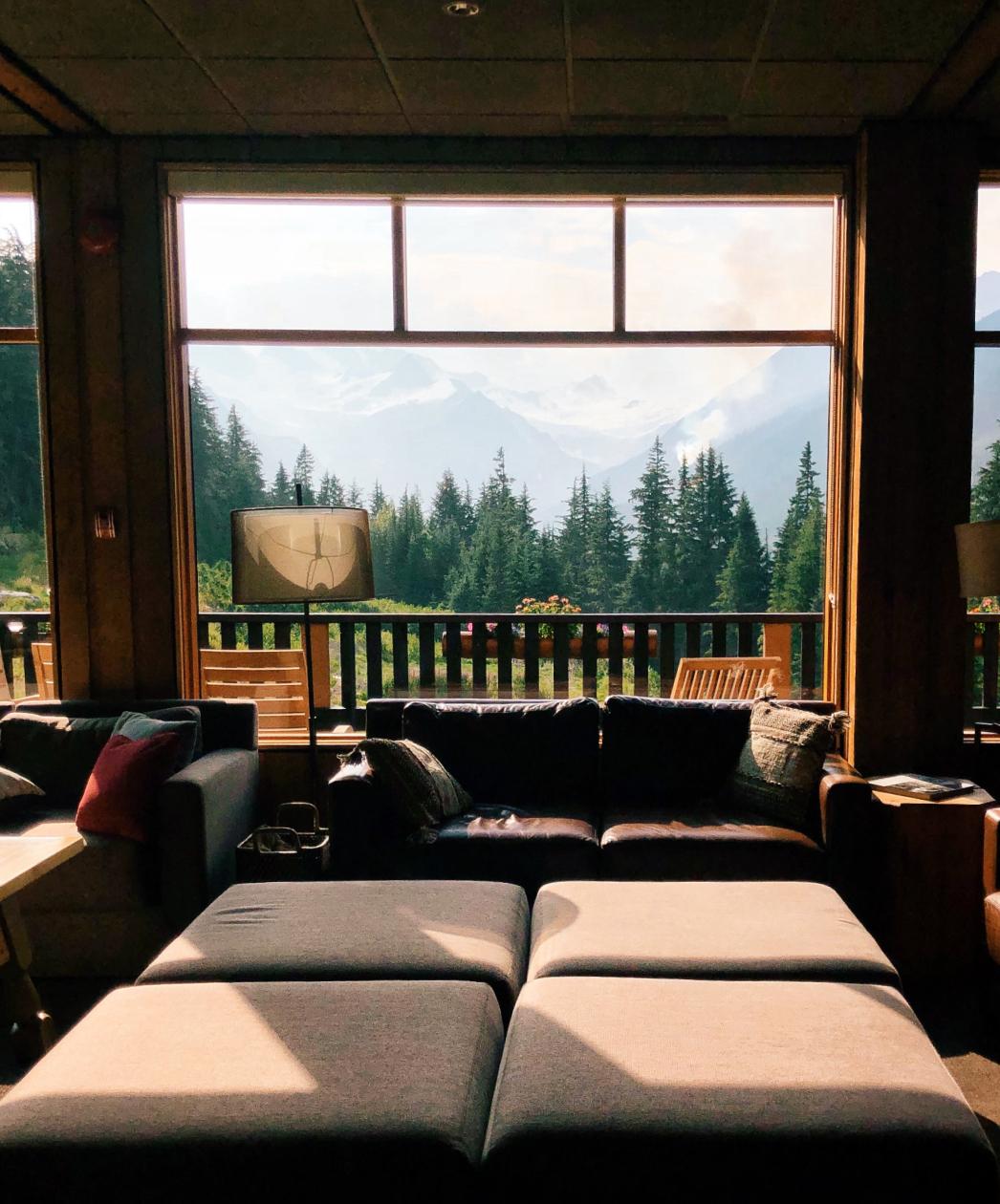 A cozy living room with sofas and ottomans facing a large window that overlooks a forest and distant mountains.