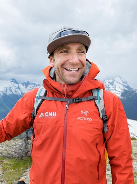 A man in an orange jacket smiling with mountains in the background