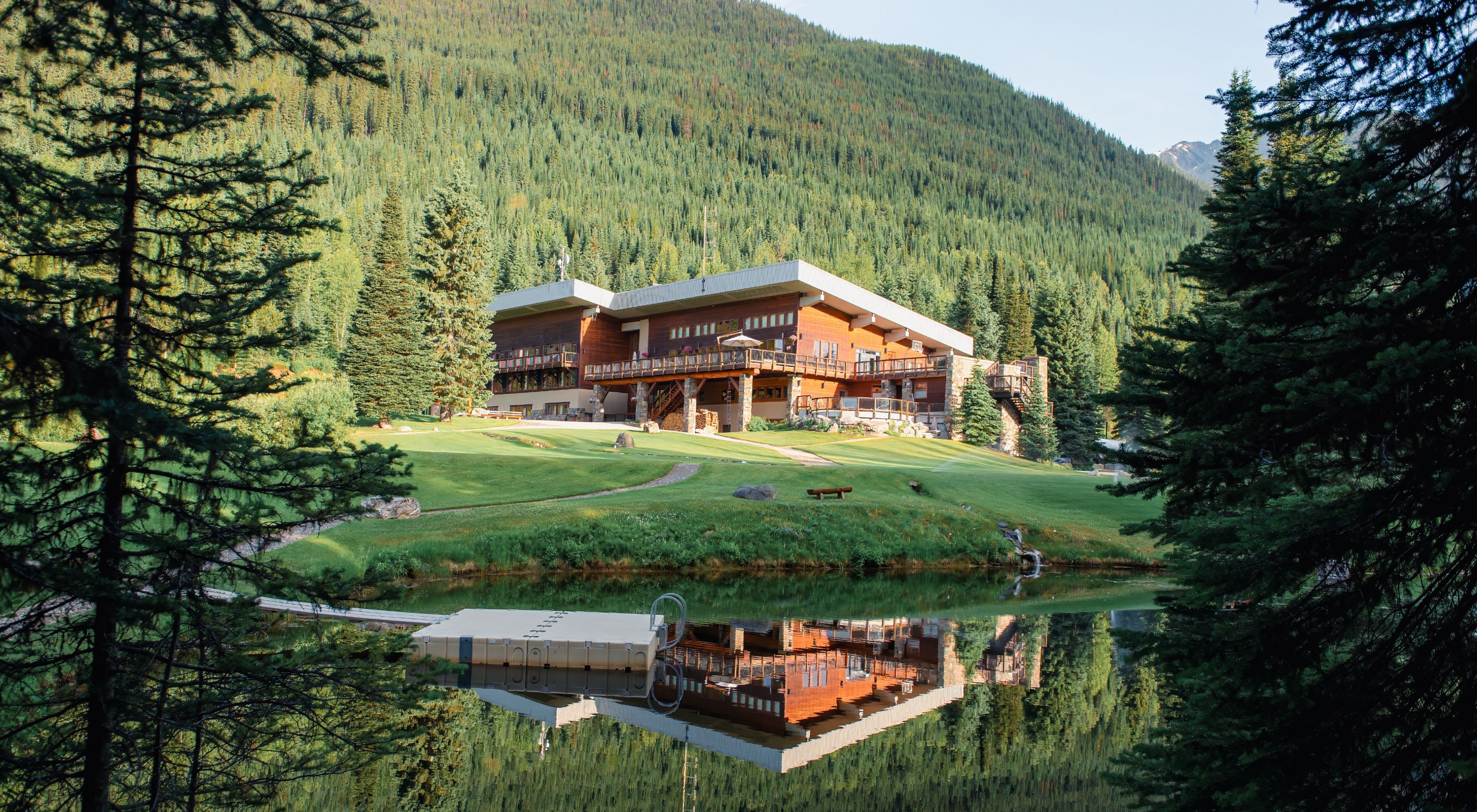 Two guests relaxing on a lodge deck surrounded by forested mountains under a bright summer sky.
