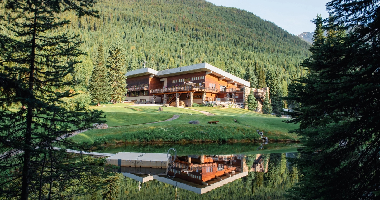 A large wooden lodge sits on a manicured grassy property beside a calm pond, surrounded by dense evergreen forest and mountains.