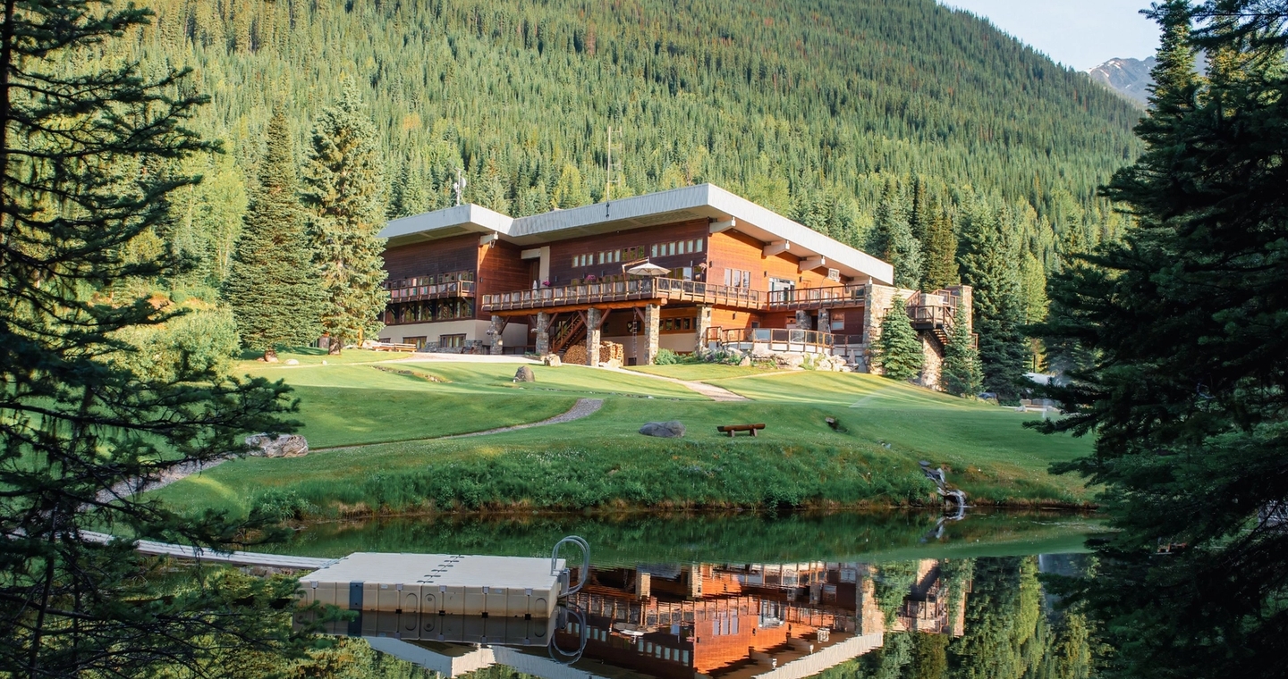 Two guests relaxing on a lodge deck surrounded by forested mountains under a bright summer sky.