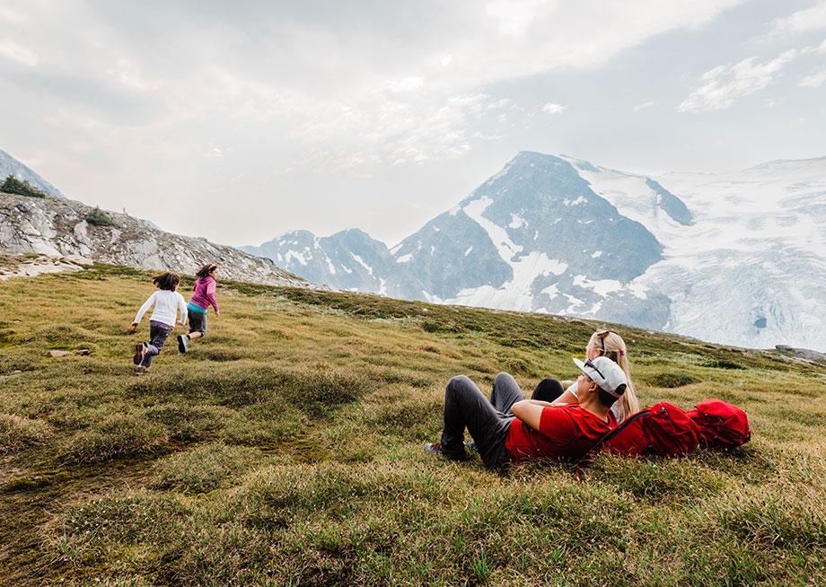 two parents lay in a meadow while their two children run around playing chase. 