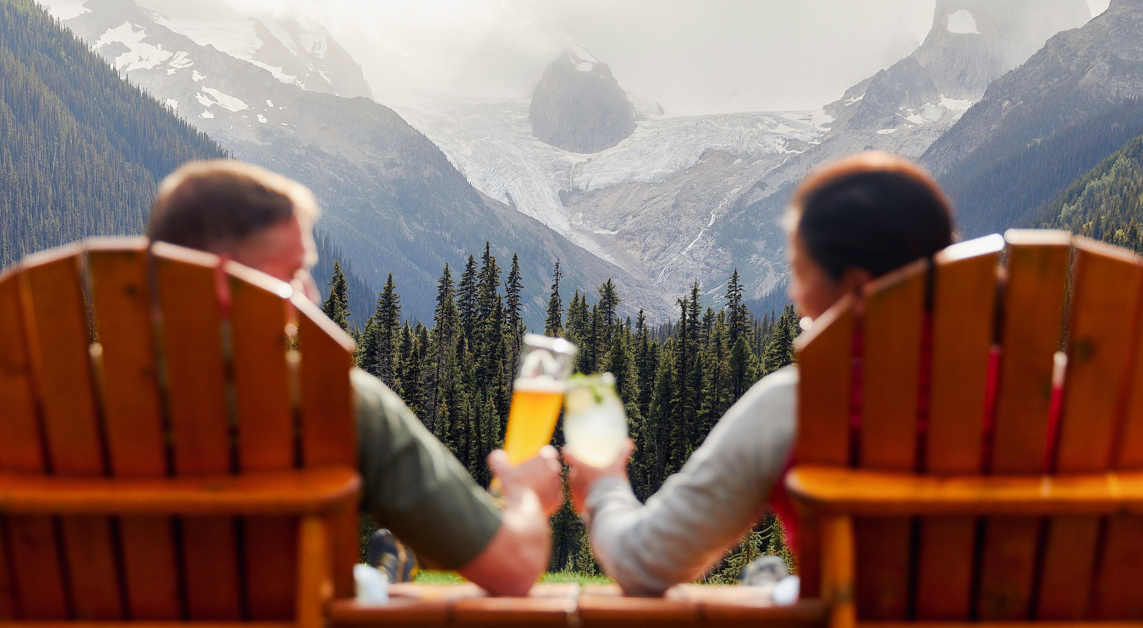 Two people sit in wooden chairs outdoors, raising drinks in a toast while facing a dramatic mountain landscape with evergreen trees, rocky peaks, and a visible glacier.