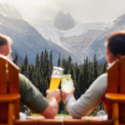 Two people sit in wooden chairs outdoors, raising drinks in a toast while facing a dramatic mountain landscape with evergreen trees, rocky peaks, and a visible glacier.