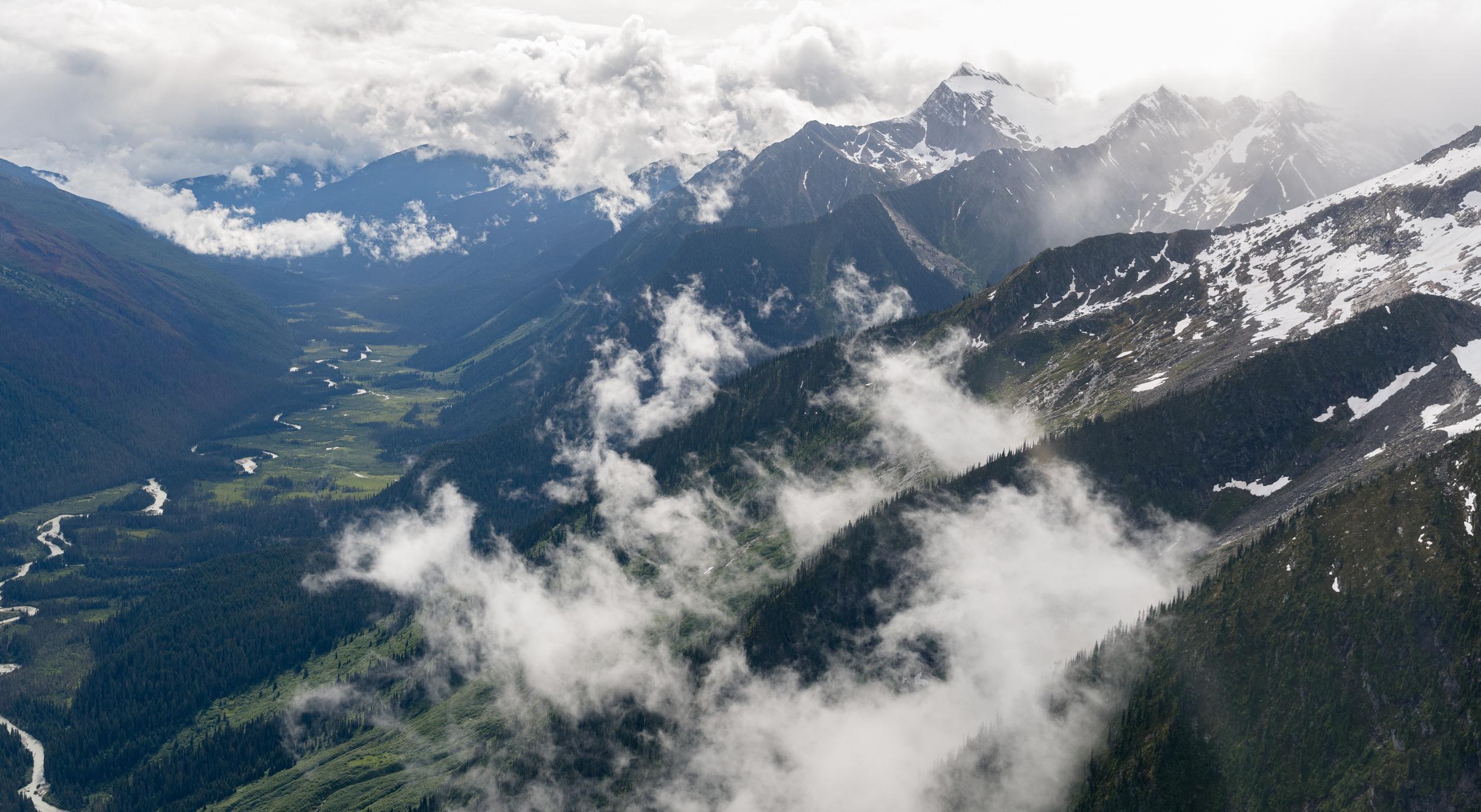 An aerial view of a deep mountain river valley surrounded by green forested slopes, low alpine clouds, and jagged snow-capped peaks during a CMH Summer Adventures backcountry tour