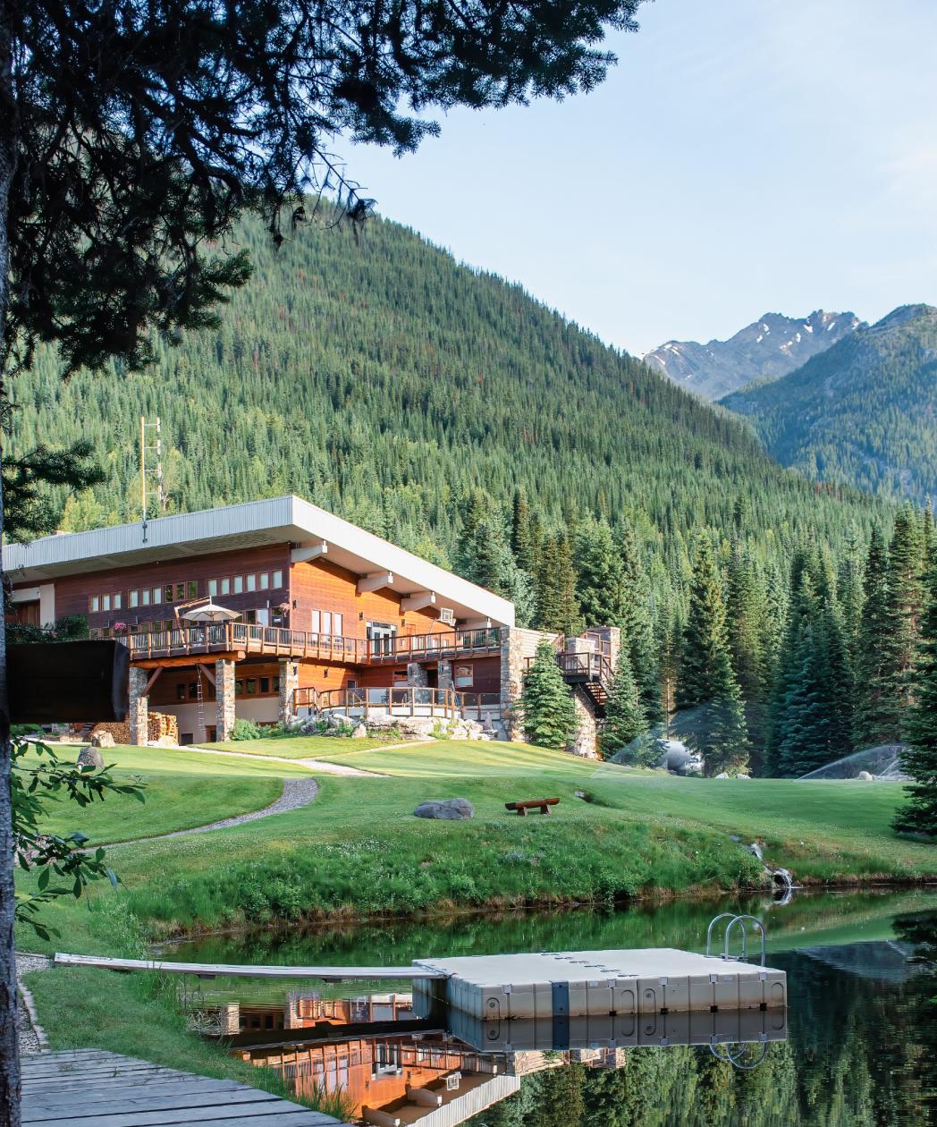 A modern wooden lodge by a small pond, surrounded by green forest and mountains under a clear sky.