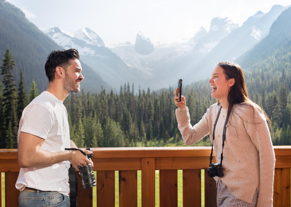 A woman taking a picture of a man with trees and mountains in the background