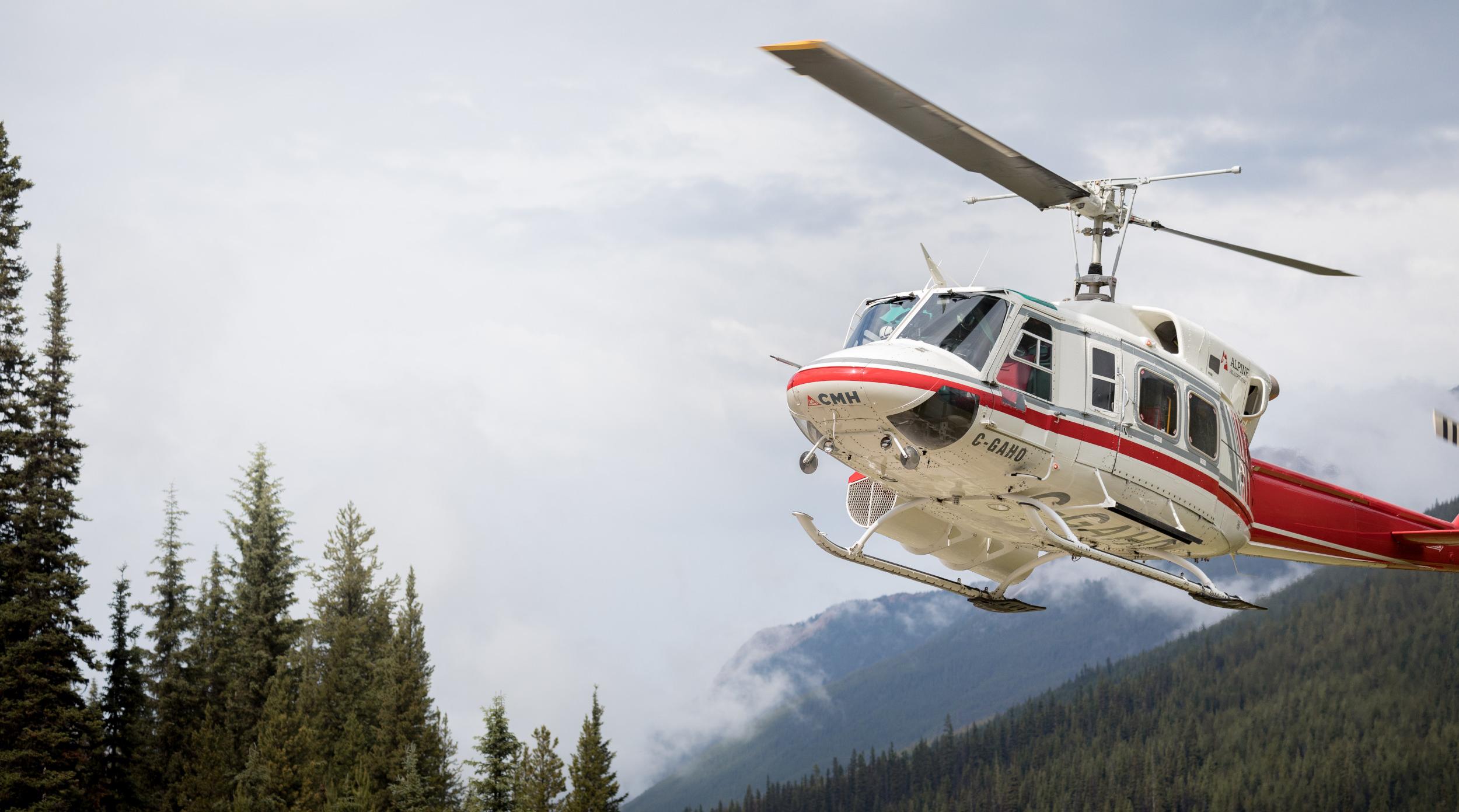 A white and red CMH Bell 212 helicopter flying low over dense evergreen pine forests and misty mountain slopes during a CMH Summer Adventures backcountry excursion