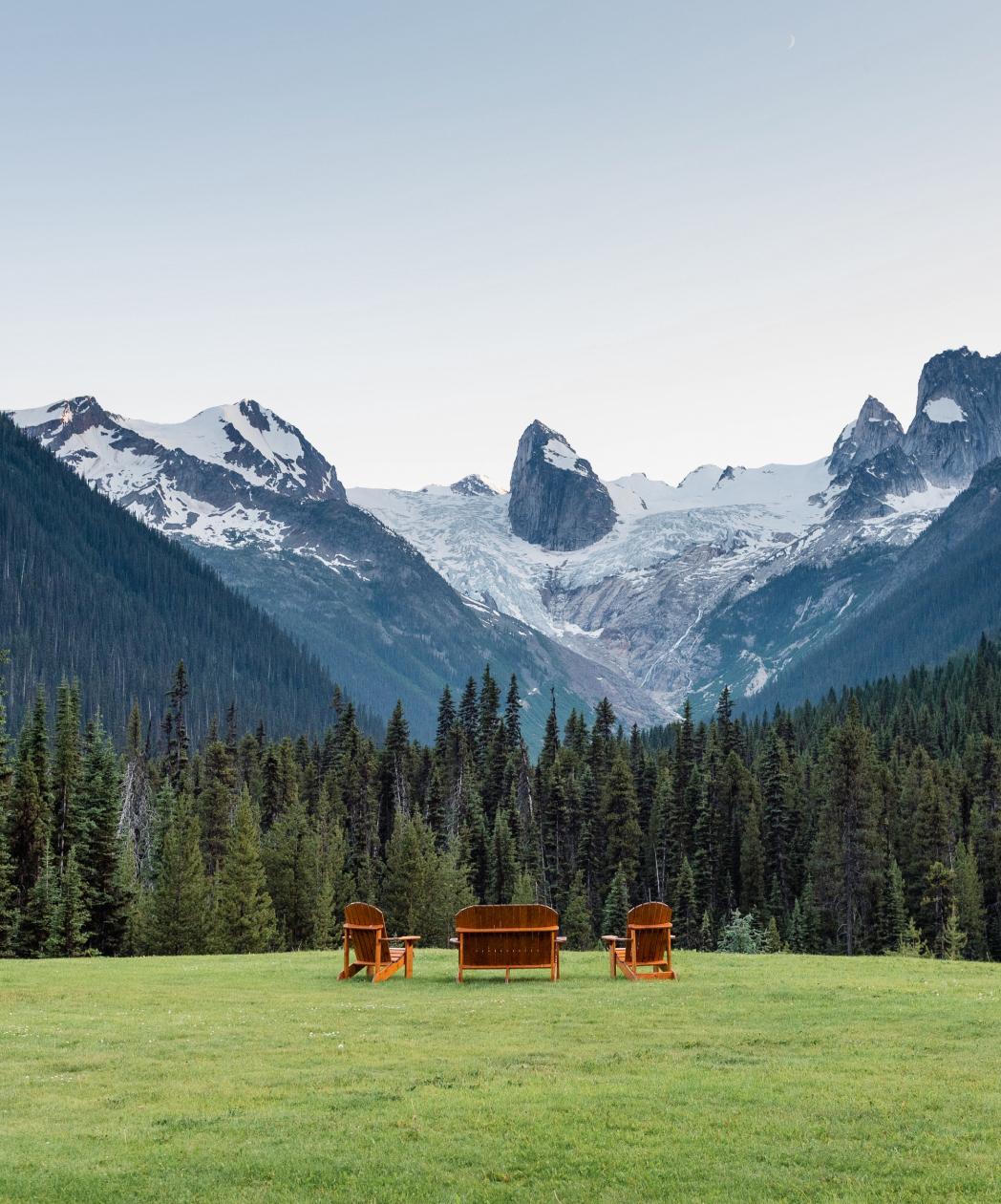 A wide, tranquil mountain landscape with three wooden Adirondack chairs arranged on a grassy field in the foreground, facing a dense forest of evergreen trees. Beyond the forest rises a dramatic range of rugged, snow-covered mountains with visible glaciers and sharp rock formations. 