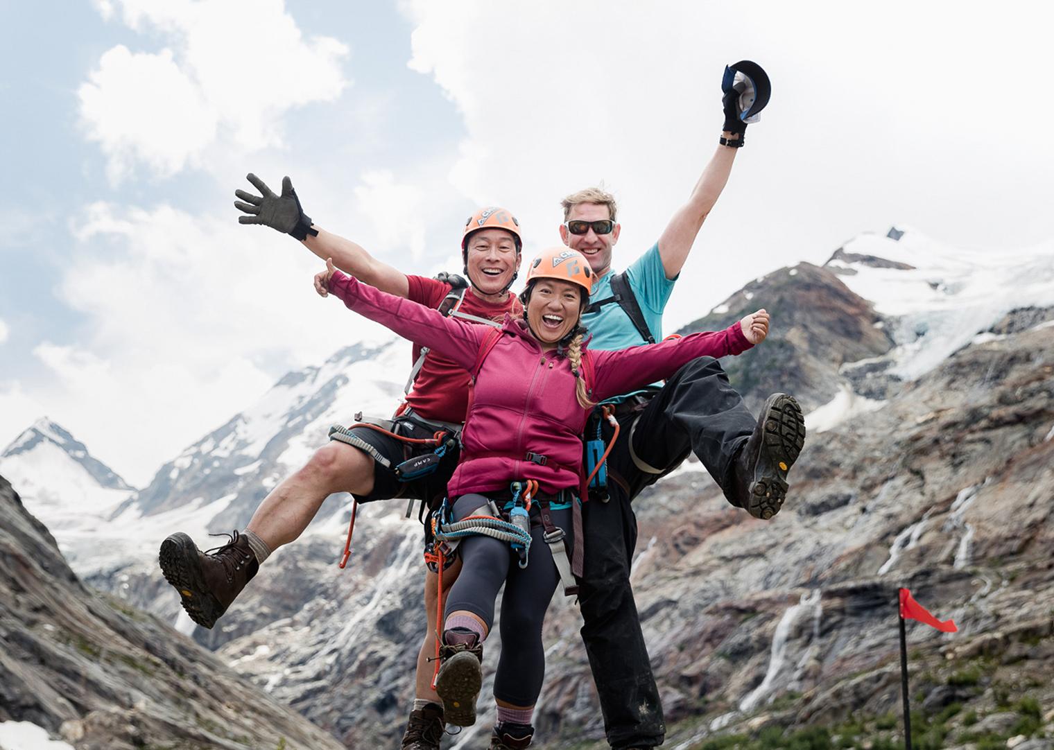 Two hikers with backpacks stand on a mountain ridge, with snow-covered peaks and a cloudy sky in the background.