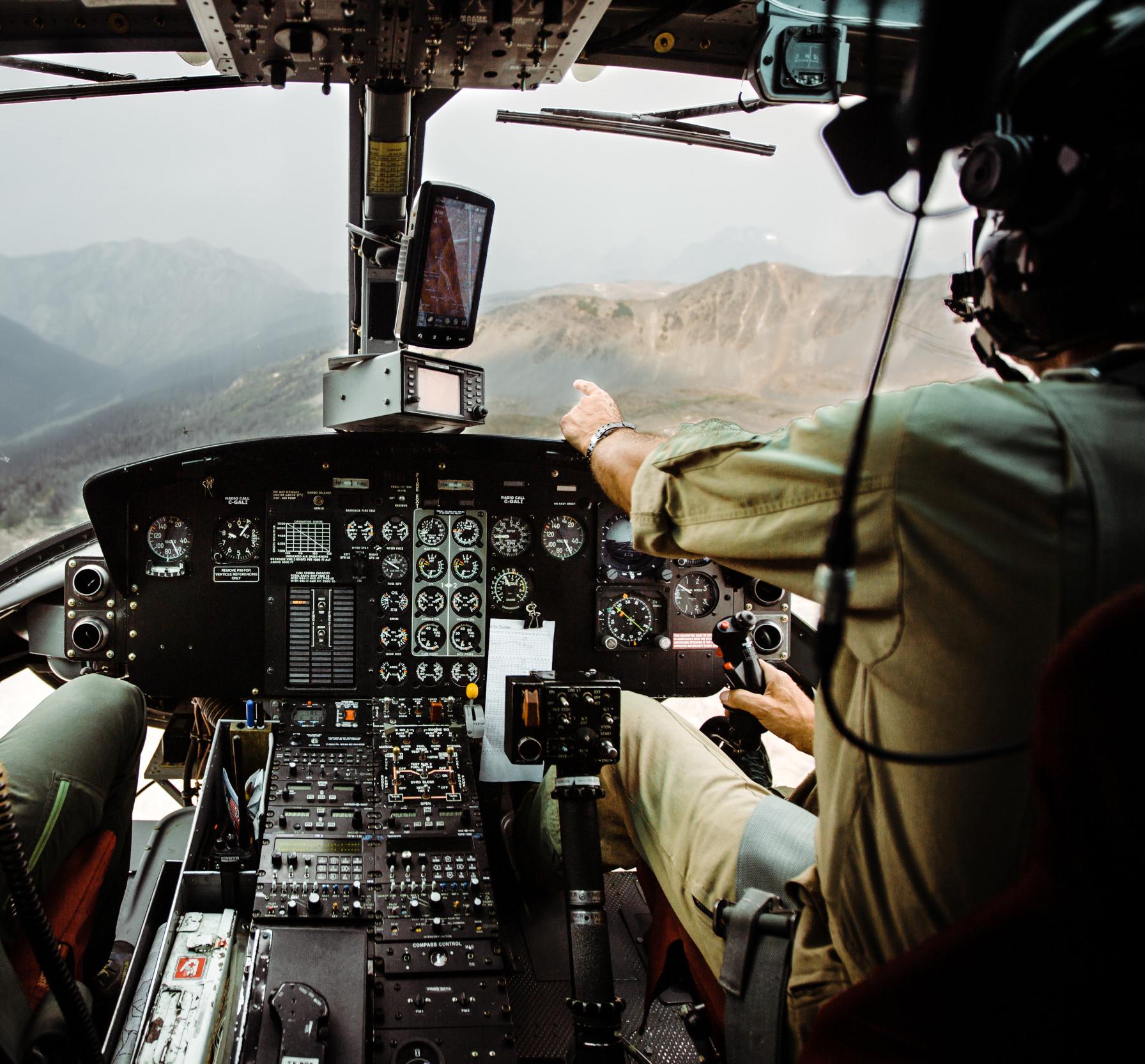 Looking into the cockpit of a helicopter, pilot pointing out the windshield at the surrounding mountains