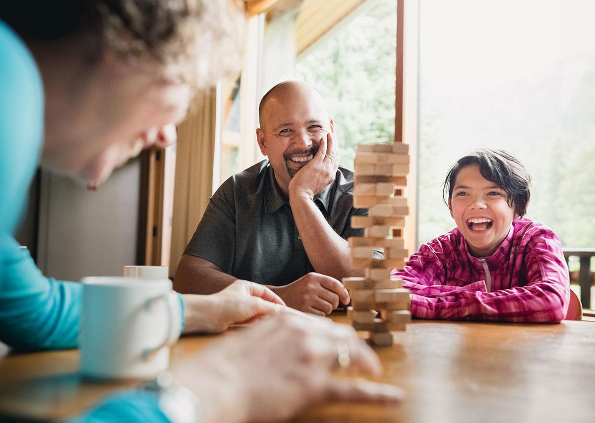 Family laughing and enjoying a game of Jenga together at a wooden table inside a cozy cabin with large windows and forest views