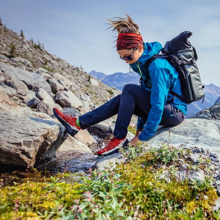 a woman in a blue jacket sitting on a rock looking down at the foliage