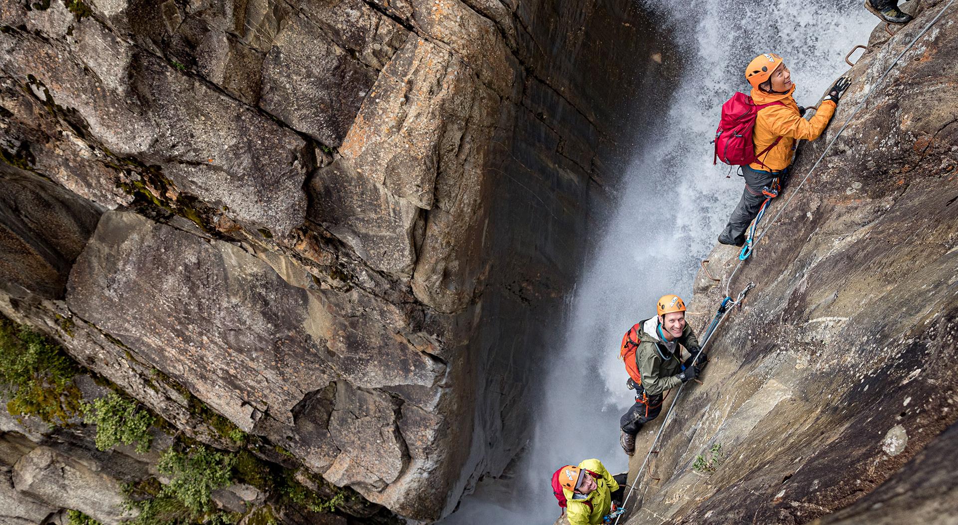 A group of guided climbers wearing safety helmets scale a steep cliff face right next to a waterfall during a CMH Summer Adventures via ferrata mountain excursion
