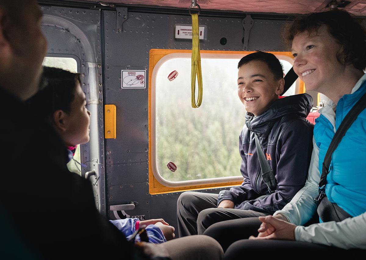 Smiling family seated inside a helicopter, looking at each other and enjoying the ride with forested scenery visible through the window.