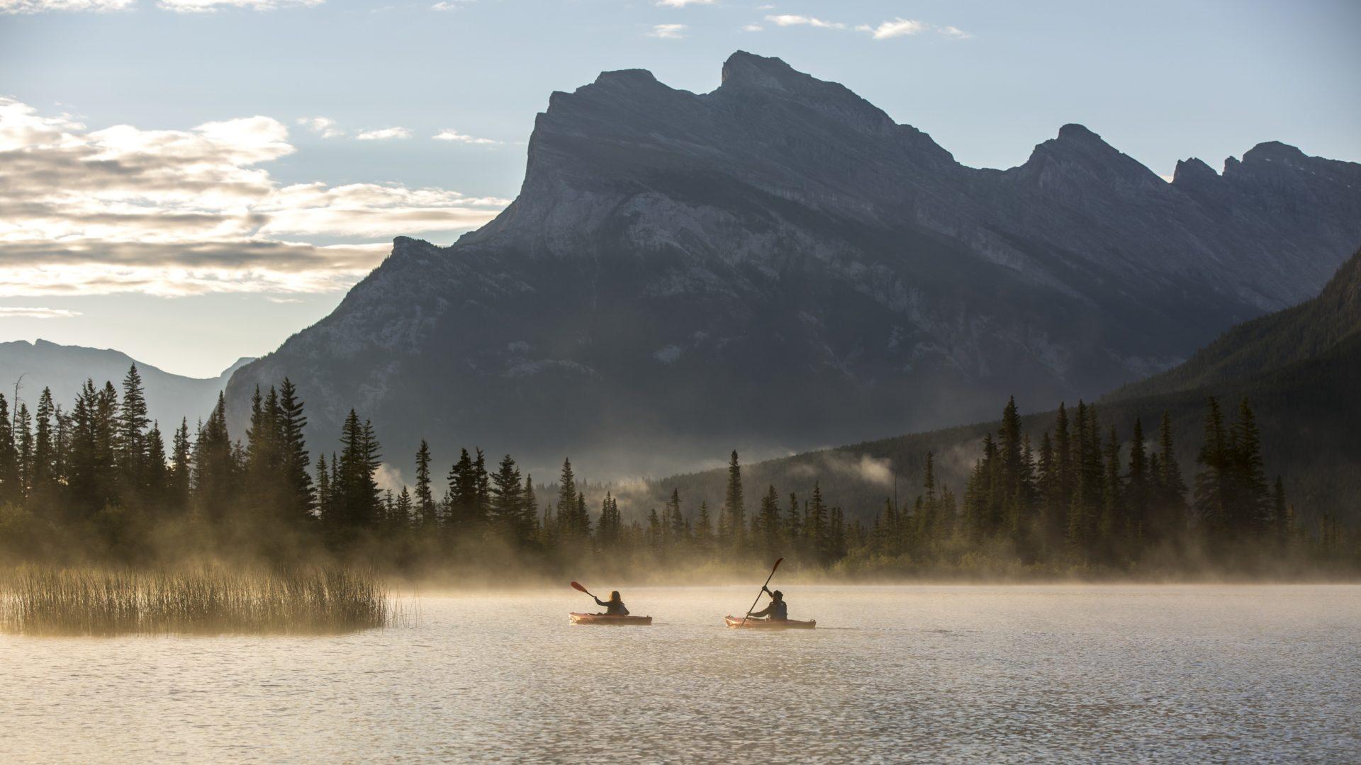 2 kayakers paddling into the sunset. A mountain range looms in the background