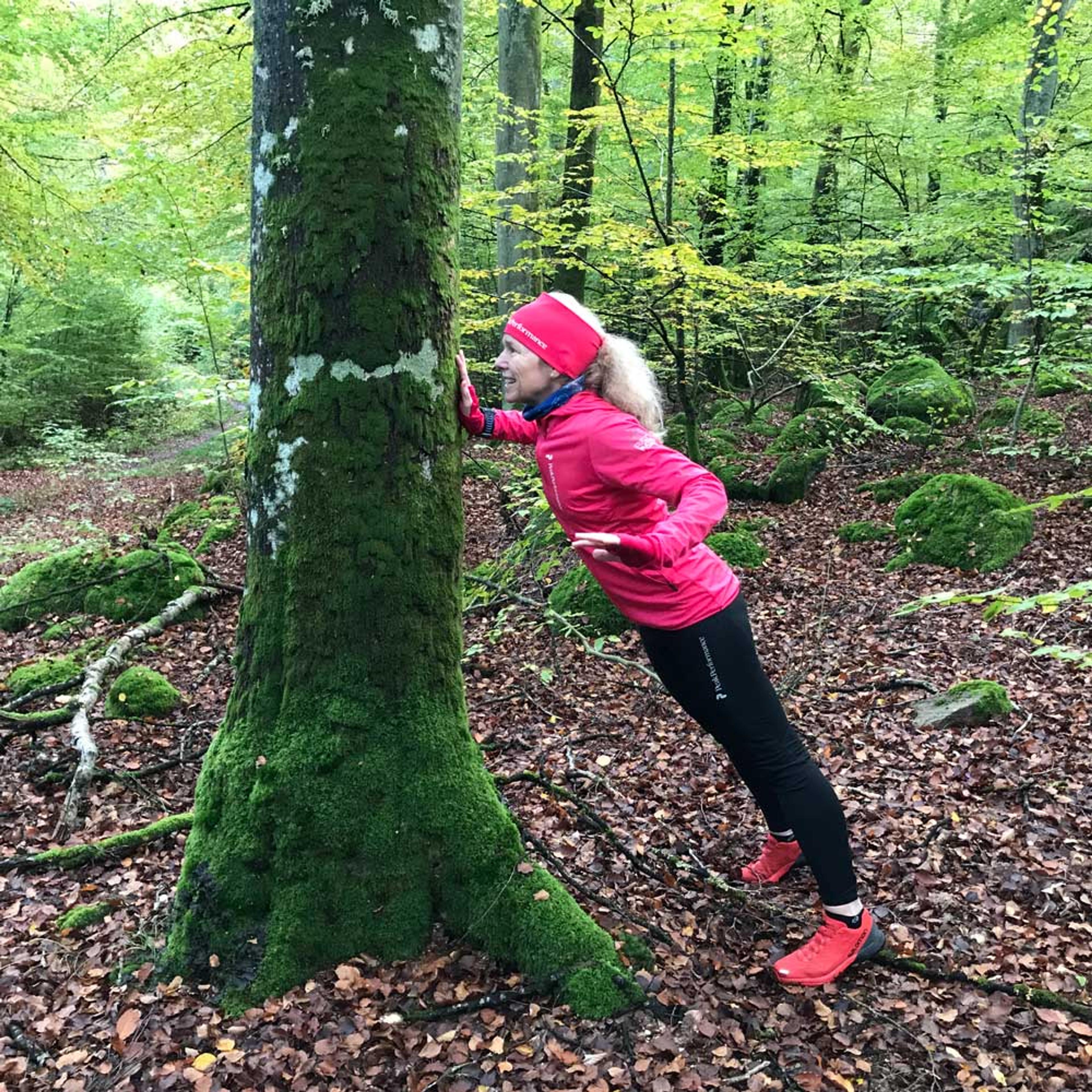 Kvinna i rosa jacka gör armhävning med en arm mot en mossig trädstam i skogen