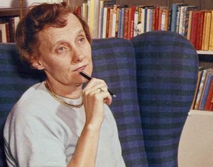 Astrid Lindgren sitter i en rutig fåtölj framför en bokhylla med en penna mot hakan