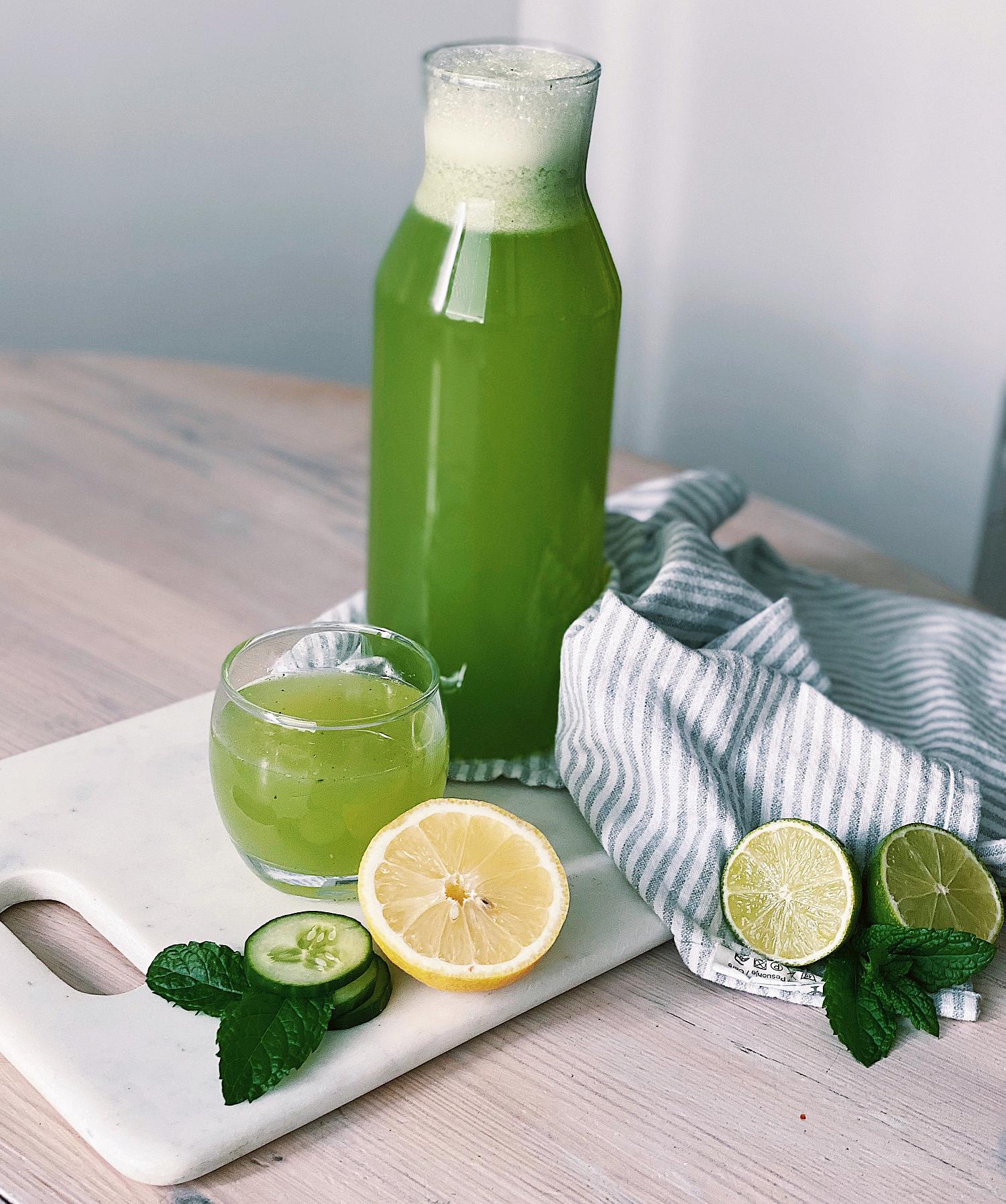Grön lemonad i en karaff och ett glas, med citron, lime, gurka och mynta på en skärbräda
