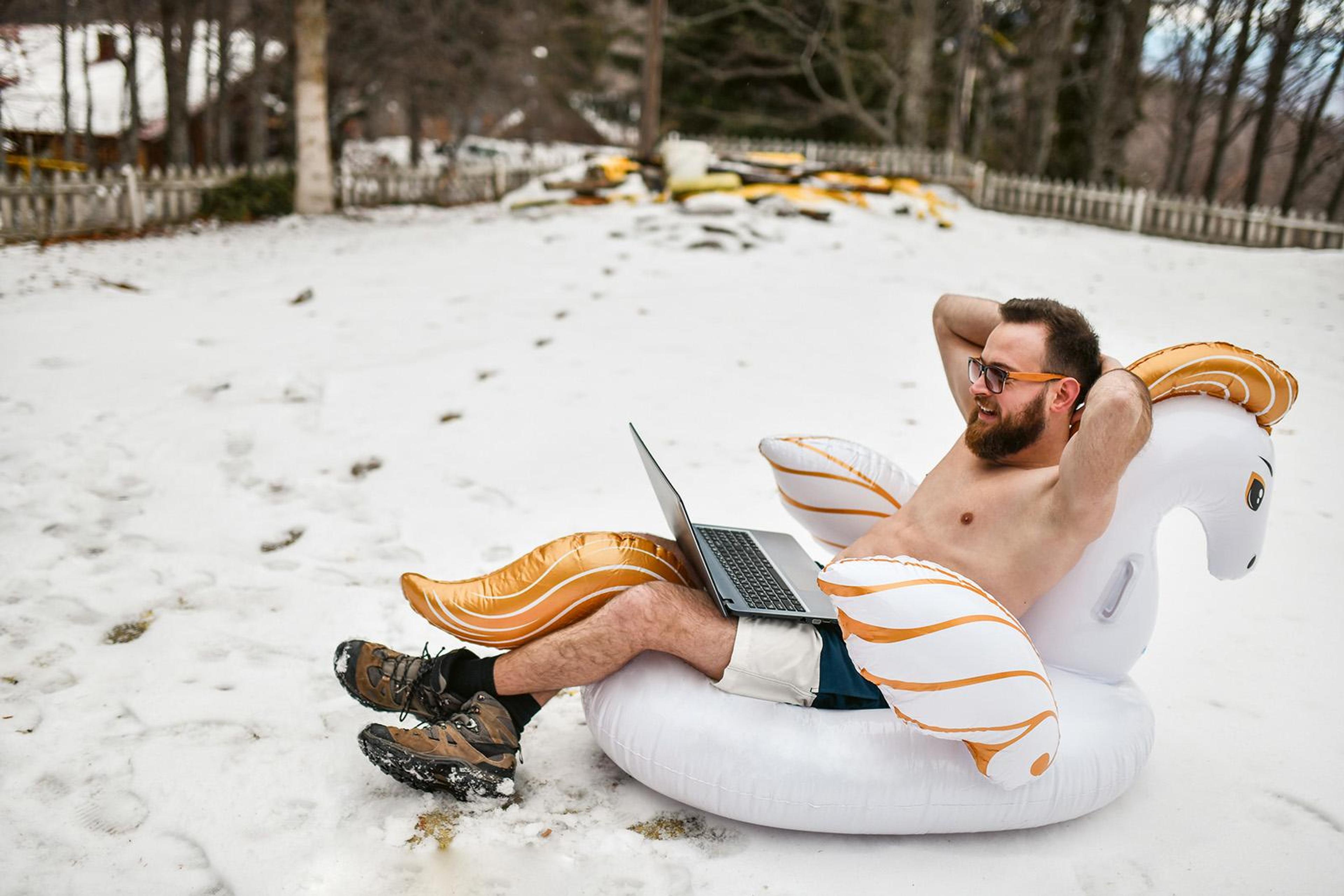 Man i bar överkropp sitter i en uppblåsbar badring i snön med en laptop i knät