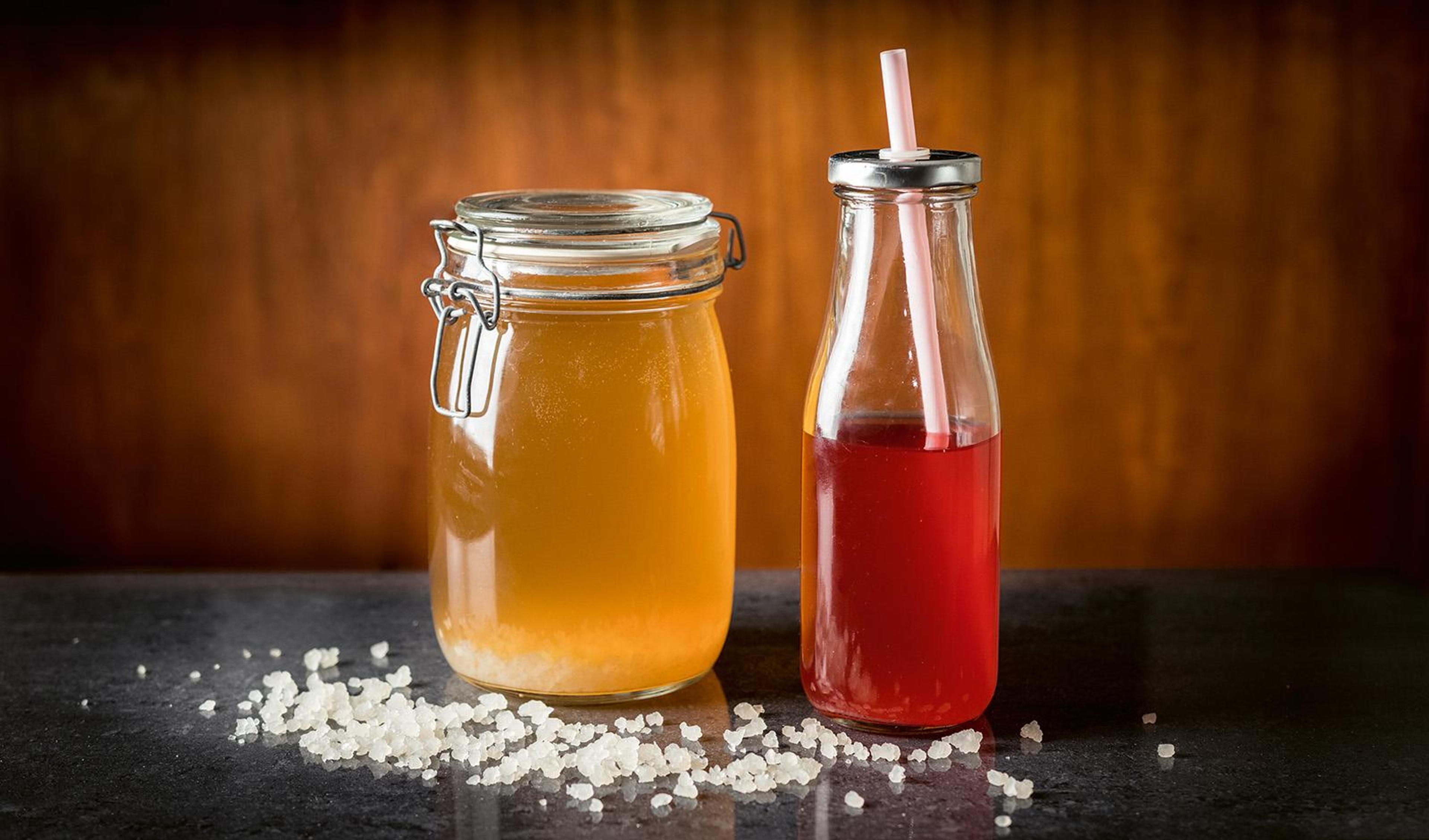 Vattenkefir i en glasburk och en flaska med sugrör, med kefirgryn utspridda framför