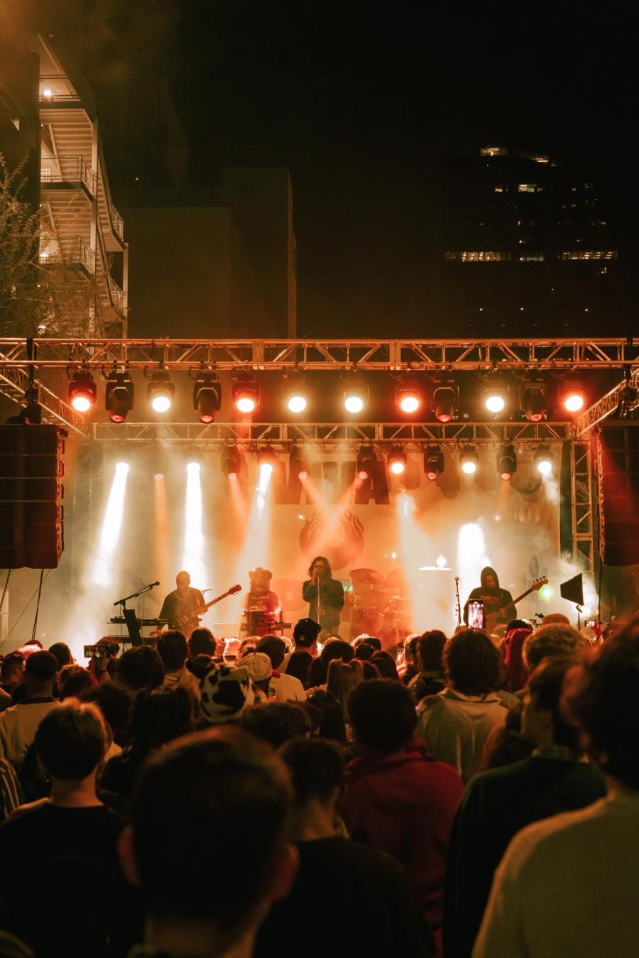 Large nighttime crowd watches a band perform on an outdoor stage lit in orange and white.