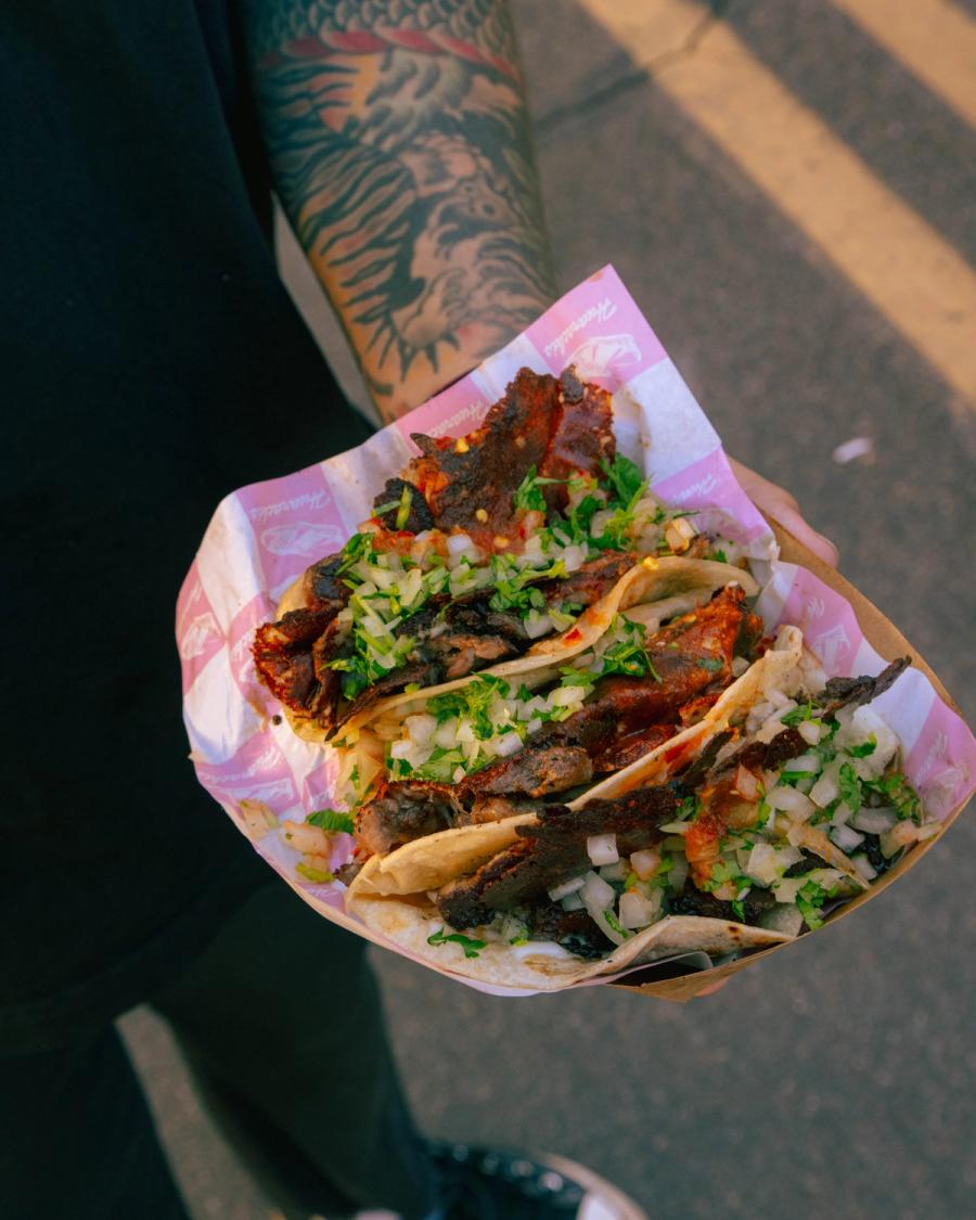 Hand holding a tray of grilled tacos topped with chopped onion and cilantro.
