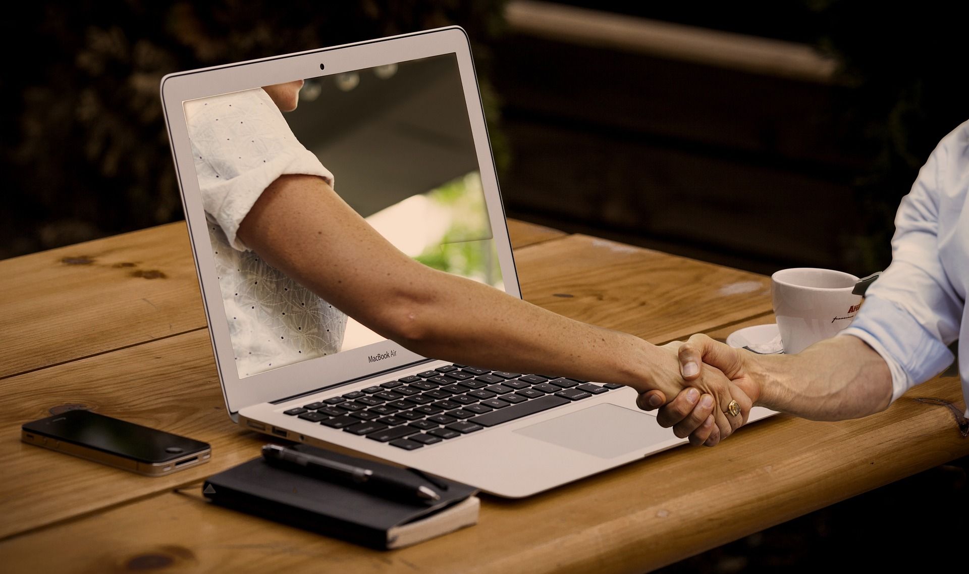 Laptop på ett bord och en hand som kommer ut från skärmen och skakar hand med en som sitter vid bordet.