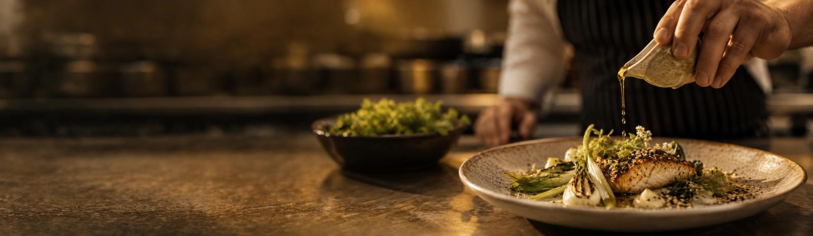 A chef in a dark kitchen drizzling sesame oil from a small glass jug over a plated dish.