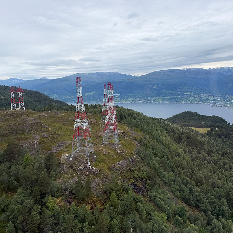 Luftfoto av flere rød- og hvitmalte kraftmaster i en skogkledd fjellside med Sognefjorden i bakgrunnen.