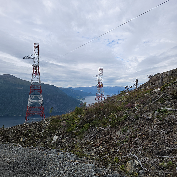 To rød- og hvitmalte spennmaster i bratt fjellterreng med Sognefjorden i bakgrunnen.