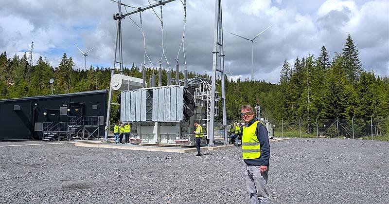 Deltakere på befaring ved Odal Vind står rundt en krafttransformator i en transformatorstasjon. I forgrunnen står en person i gul refleksvest, mens vindturbiner og skog danner bakgrunnen.