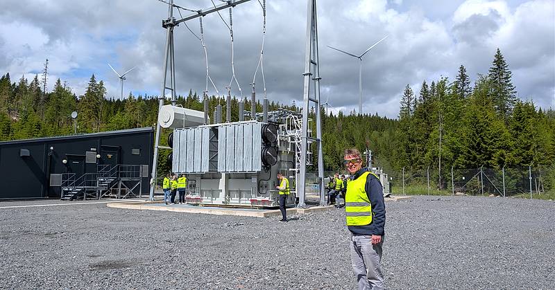 Deltakere på befaring ved Odal Vind står rundt en krafttransformator i en transformatorstasjon. I forgrunnen står en person i gul refleksvest, mens vindturbiner og skog danner bakgrunnen.