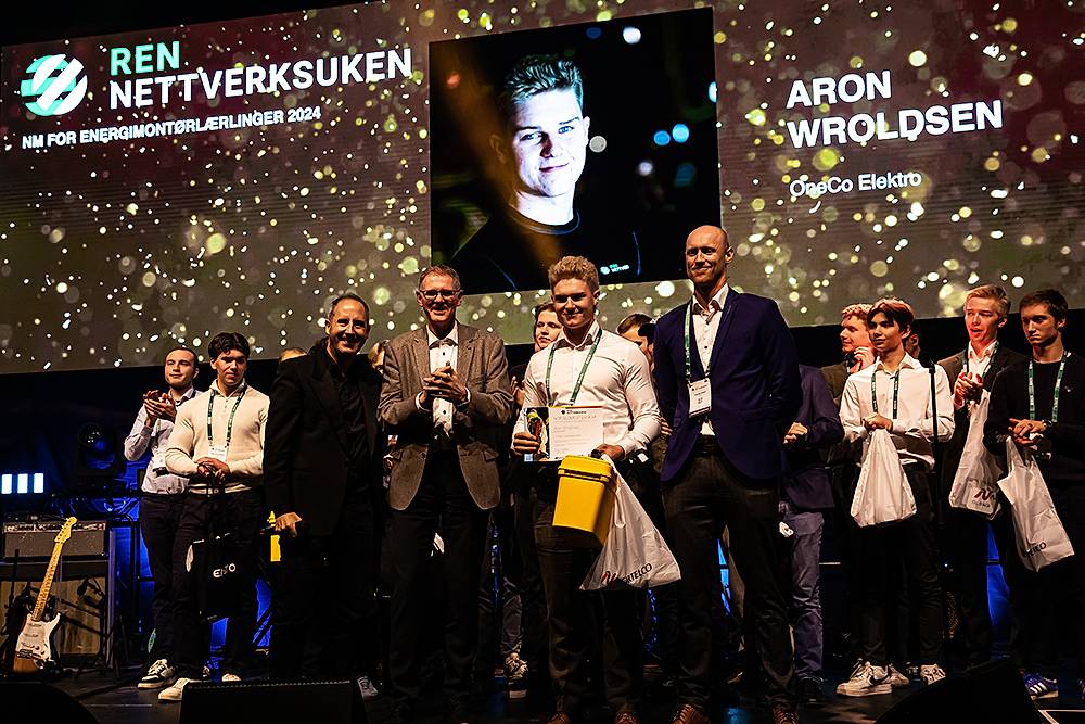 Bilde fra prisutdeling til NM for energimontørlærlinger 2024. Bildet viser konferansier Harald Rønneberg, fagansvarlig NM Kai Solum, norgesmester Aron Wroldsen og hovedansvarlig Andreas Solum fremme på scenen. De øvrige NM-deltakerne står bak.