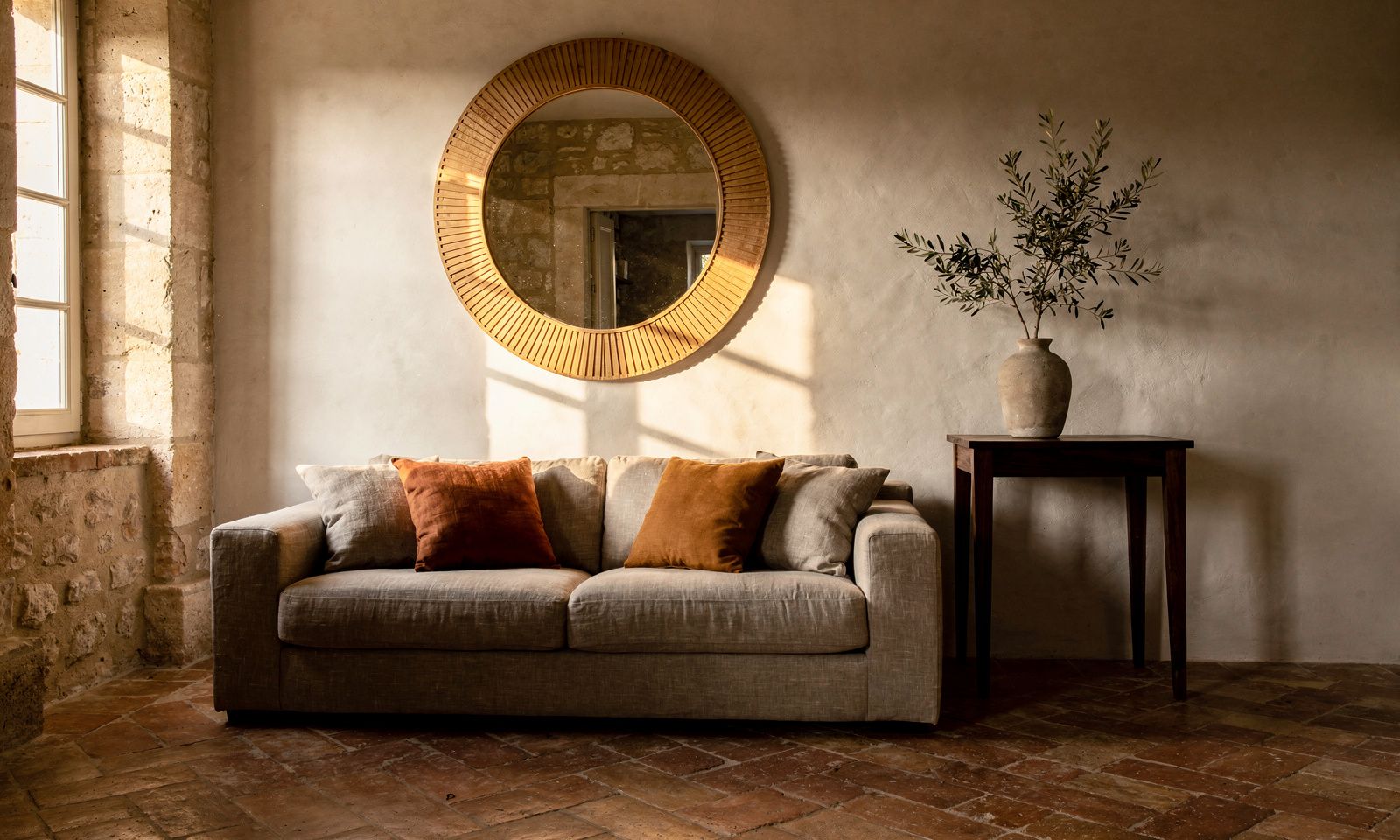 Living room in a house in Châteauneuf-Grasse with a large round mirror in a pale oak frame, lime-plaster walls and soft late-afternoon light